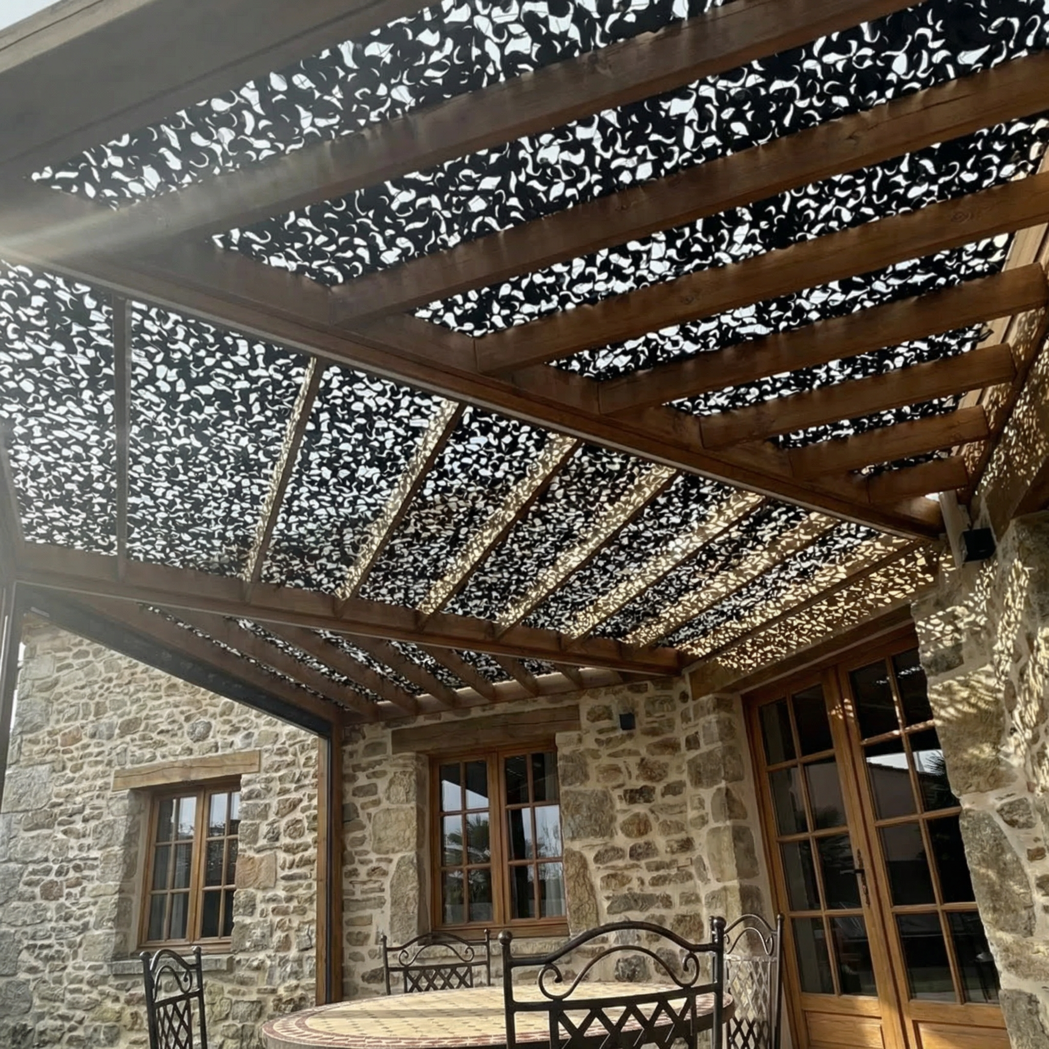 Filet de camouflage noir renforcé ombrageant une terrasse en pierre avec structure en bois.